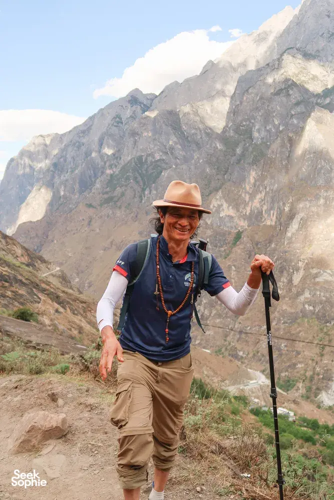 Smiling trekker hiking along a dirt path in Tiger Leaping Gorge, Lijiang, with steep granite peaks towering behind