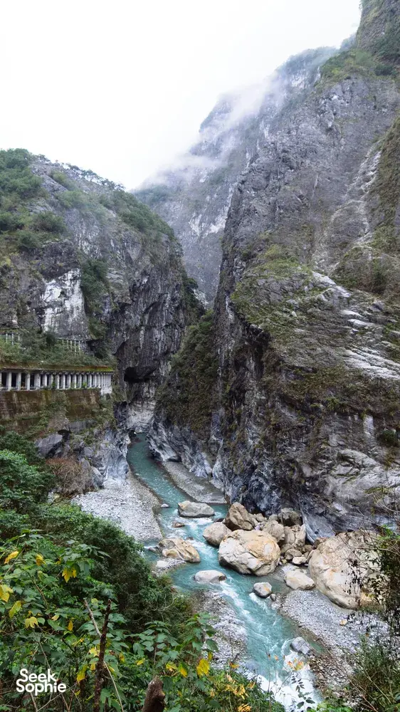 Narrow mountain gorge trail with a clear blue stream cutting between steep rock cliffs and a riverside bridge in China