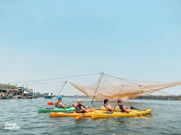 People in kayaks with life jackets paddle along the river in Hoi An, passing moored boats and netted harbour views