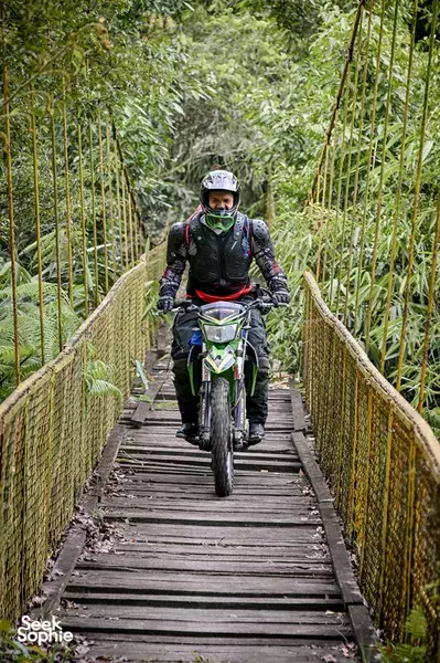 Motorbike rider crosses a bamboo suspension bridge in dense jungle on an Indonesia motorcycle tour, with a dirt road ahead