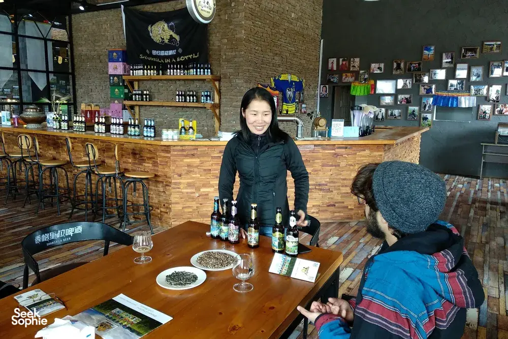 Smiling host pours while visitors sit at a wooden table with Tibetan beer bottles in a cosy brewery bar in Shangri-La