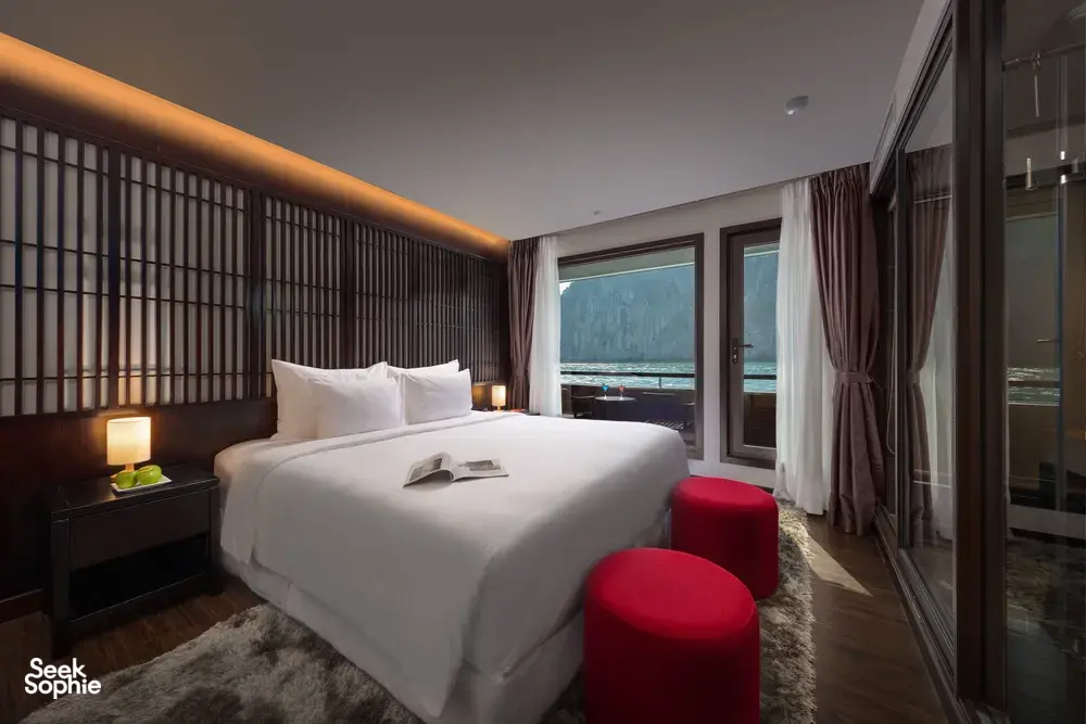 Oasis Bay Party Cruiser cabin with a made-up bed, red ottoman seating and sea-view windows overlooking karst scenery on the Halong Bay