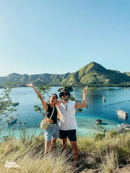 Two hikers posing with arms wide at a scenic coastal overlook, calm blue bay and boats below with green hills