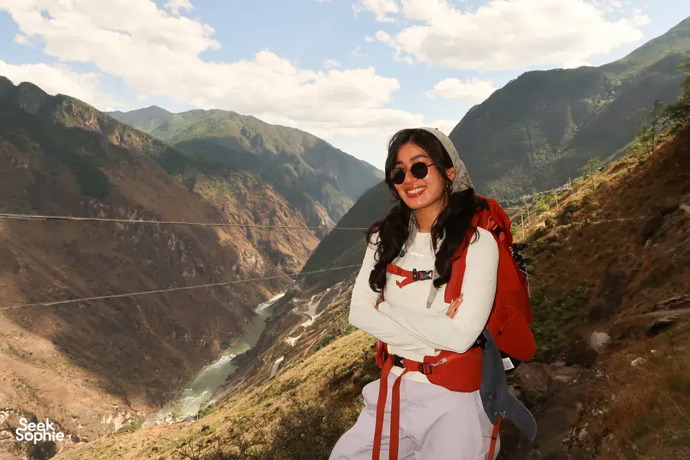 Smiling hiker in sunglasses with a red backpack on the cliff path above Tiger Leaping Gorge in Lijiang with The Bivou Lijiang