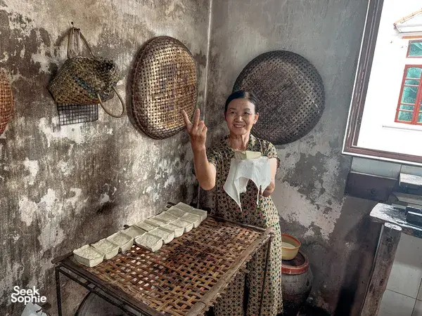 Ngoc from Bamboo Life pressing tofu on a bamboo lattice in a Hanoi kitchen, with woven hats hanging on the wall