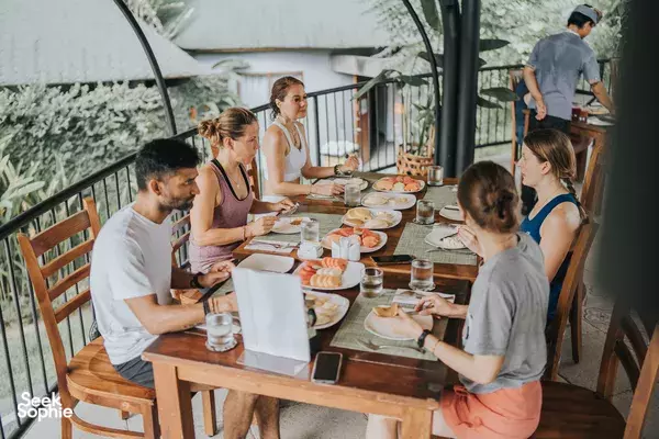 Group seated at a long wooden table eating together outdoors, with plates and drinks on the table at an Indonesian yoga retreat