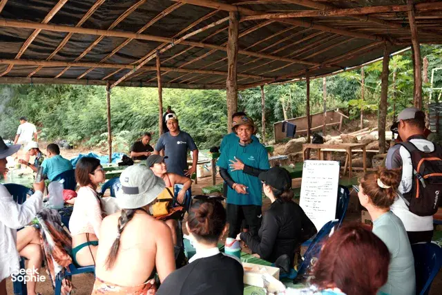 Groups seated on plastic chairs for a coral nursery briefing while guides gesture under a shaded pavilion on Komodo Island