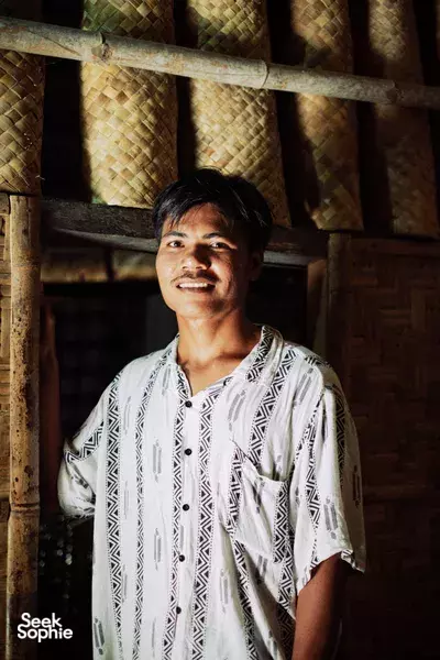 Local guide smiling in a doorway of a traditional thatched house on Lombok, wearing a patterned shirt