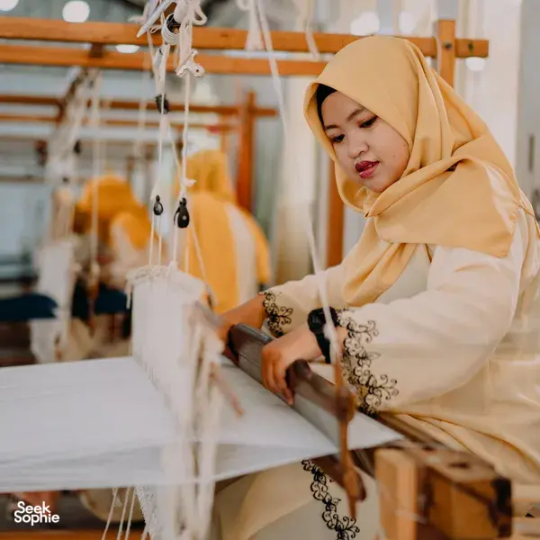 Headdress-clad artisan stitching threads on a white loom at Tanoti Crafts weaving workshop in Kuching, Sarawak