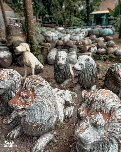Weathered stone lion statues along a garden path, with greenery and other cast sculptures around a craft village, Yogyakarta