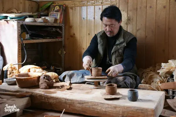 Potter at a workbench carving and shaping small cups from clay in a Shangri-La Tibetan home workshop during pottery time