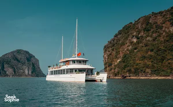 White sailing cruise yacht gliding on calm water with steep jungle-covered cliffs and a second vessel nearby, Halong Bay