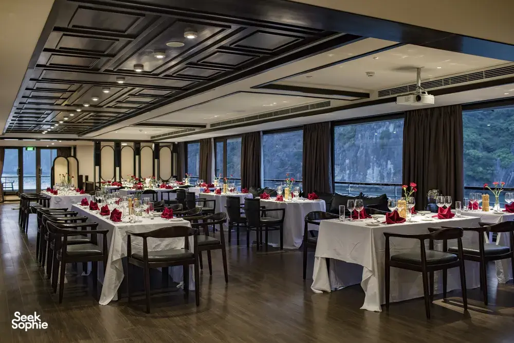 Set dining tables with red napkins and flowers inside a party cruise lounge, looking out over Halong Bay