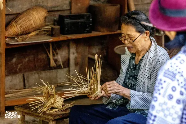 Woman sits in a rustic kitchen workshop, braiding dried straw or pine needles for ingredients in a Mae Kampong forage-to-table class