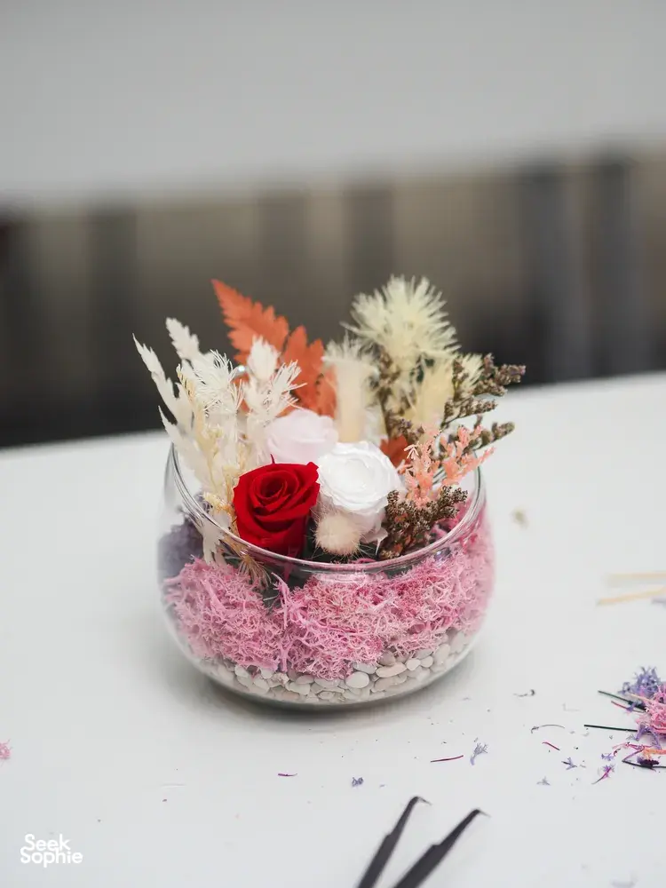 Preserved floral bowl terrarium with a red rose and orange blooms on pink moss, surrounded by petals on a worktable in Singapore