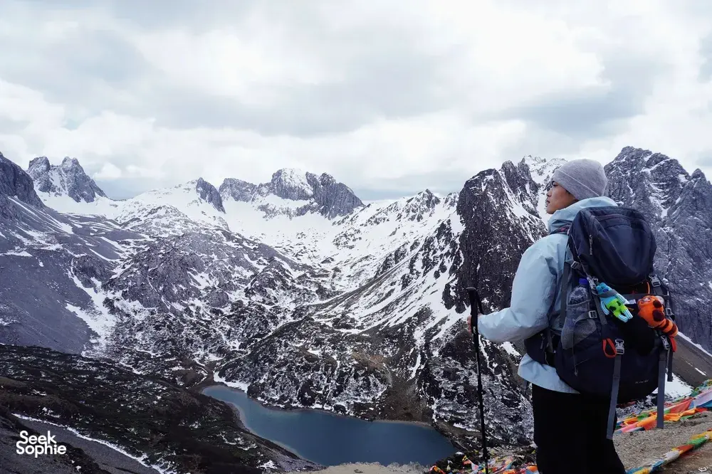 Backpacker with trekking poles overlooks a snowy valley and turquoise water in Shangri-La, hiking at high altitude