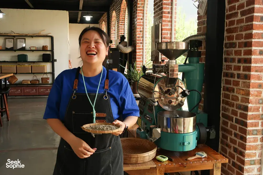 Host-led specialty coffee workshop at Seeker Yunnan Coffee Estate, Lijiang: woman holds a tray of roasted beans beside a roaster
