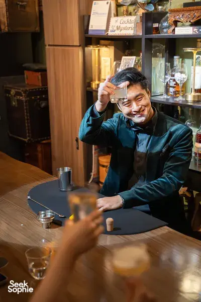 Magician in blue jacket smiling as he lifts a small item near glassware at a Magic Workshop in Singapore at workshops in Singapore