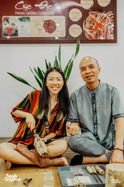 Two guests seated in a Hanoi agarwood workshop, smiling beside carved agarwood pieces on a wooden table
