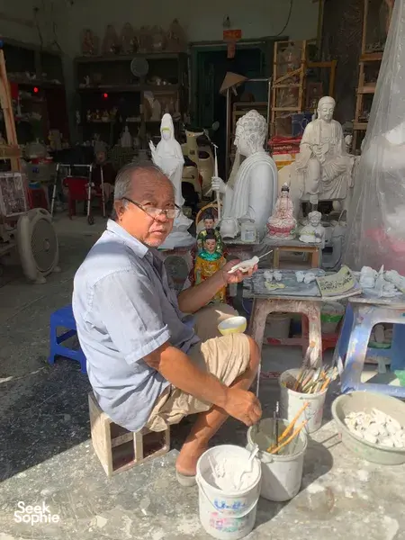 Elderly artisan painting small figurines and surrounded by white Buddha sculptures at a craft shop in Ho Chi Minh City