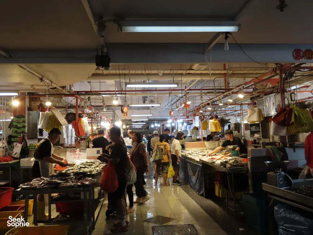 Lorong pasar makanan laut yang sibuk di Singapura dengan gerai penjaja dan pengunjung, menunjukkan dari mana ikan Kanton diperoleh dalam lawatan