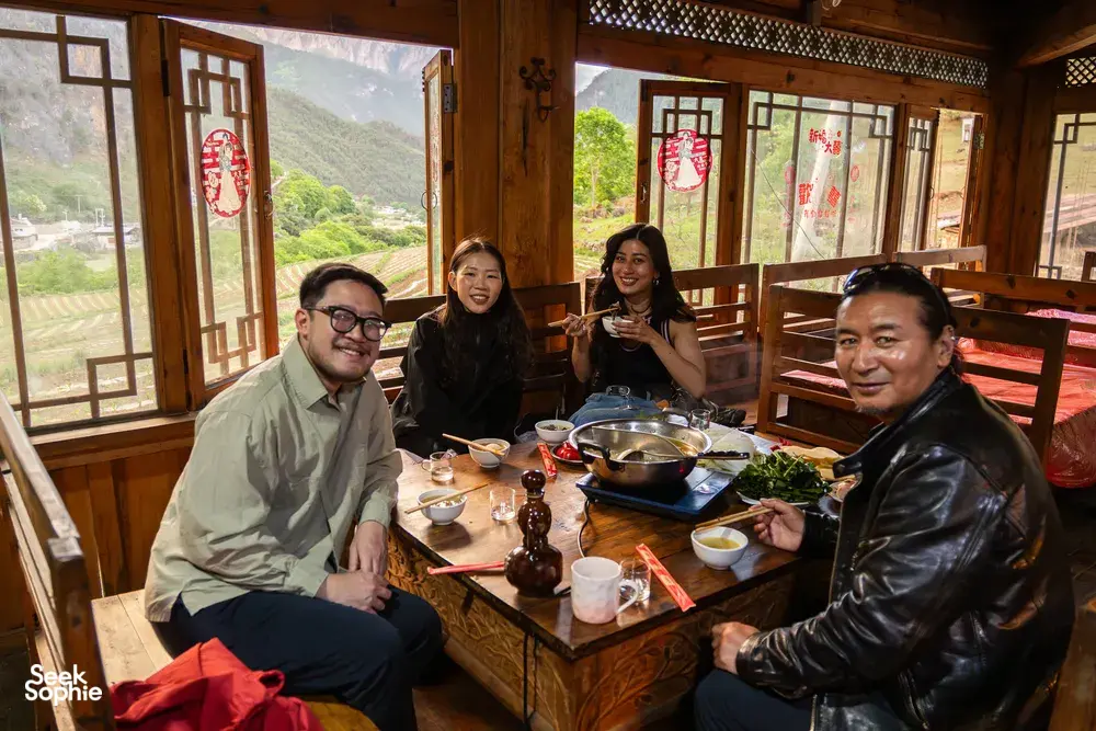 Group sharing a meal inside a traditional dining room in Shangri-La, with large windows framing a green mountain valley