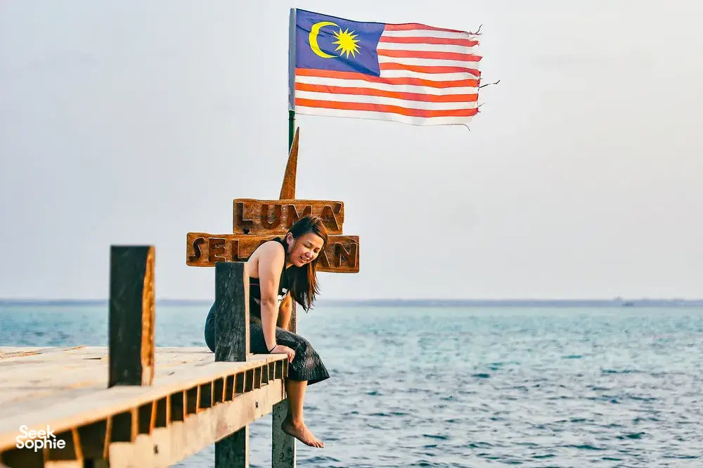 Traveller seated on a wooden jetty with Malaysian flag at Luma Selakan sea lodge over the water in Semporna