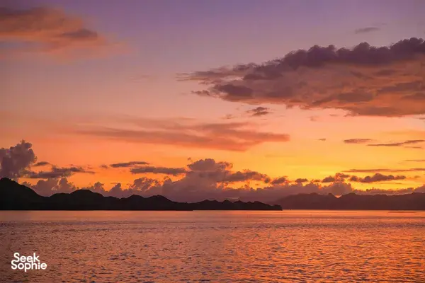 Sunset over calm sea with dark island silhouettes on the horizon, warm orange clouds reflecting on the water at dusk