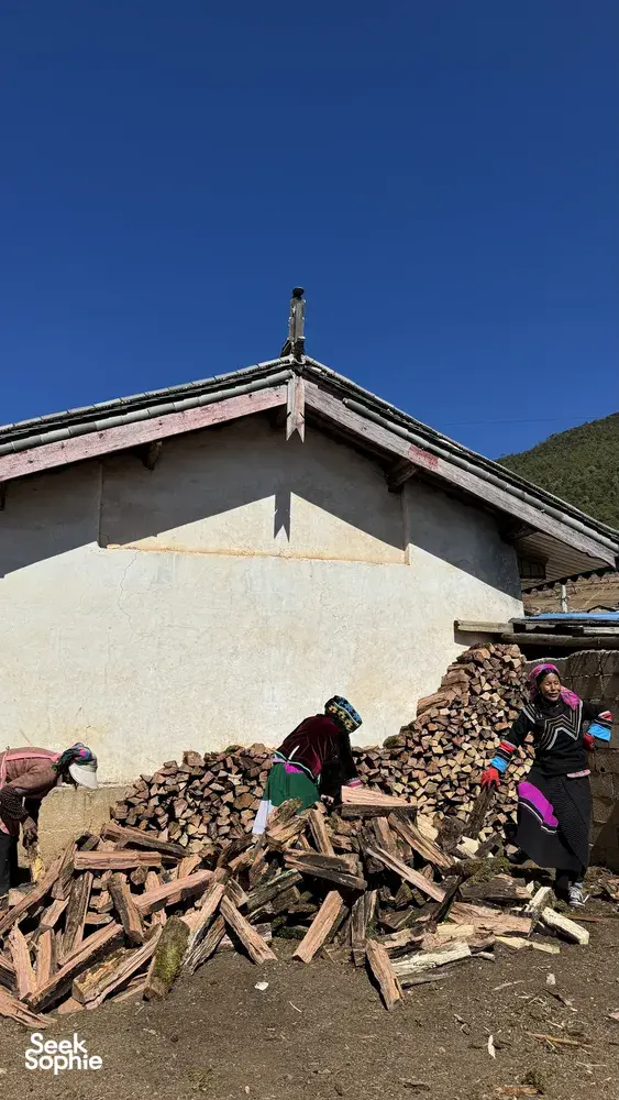 People in colourful traditional clothing stand by a rural house and stacked firewood in a Lijiang Yi & Naxi village