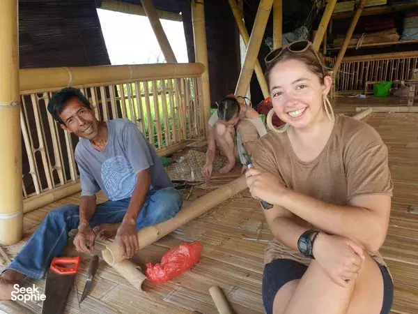Smiling workshop participant holds a bamboo pole while a man works in the background and others fit pieces inside a Bali bamboo build