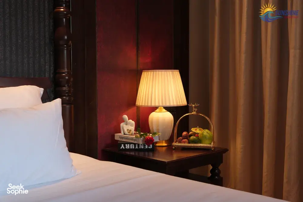 Sunshine Golden Cruises cabin bedside table with an illuminated lamp, fruit tray and bundled bedside linens against dark wood walls