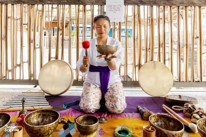 ADDA Yoga sound healer holding a brass singing bowl with resting bowls on a mat, bamboo studio session in Canggu