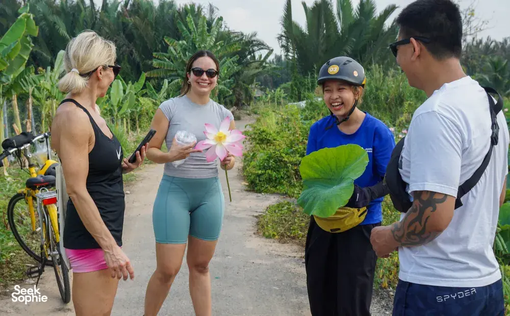 Cyclists on a narrow dirt track in lush greenery, one holding a pink lotus, during a bike tour of Mekong Island near Ho Chi Minh City