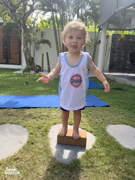 Young child balances on a small square prop on a yoga mat while participating in a playful wellness session in a Bali villa, Canggu