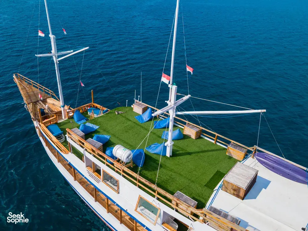 Top view of a wooden Komodo boat deck in Labuan Bajo, with green matting, flags on the masts, and calm blue sea