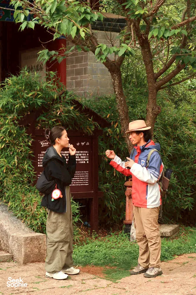 Local guide speaking with travellers beside a stone wall and bamboo garden in Lijiang’s old town courtyard walk