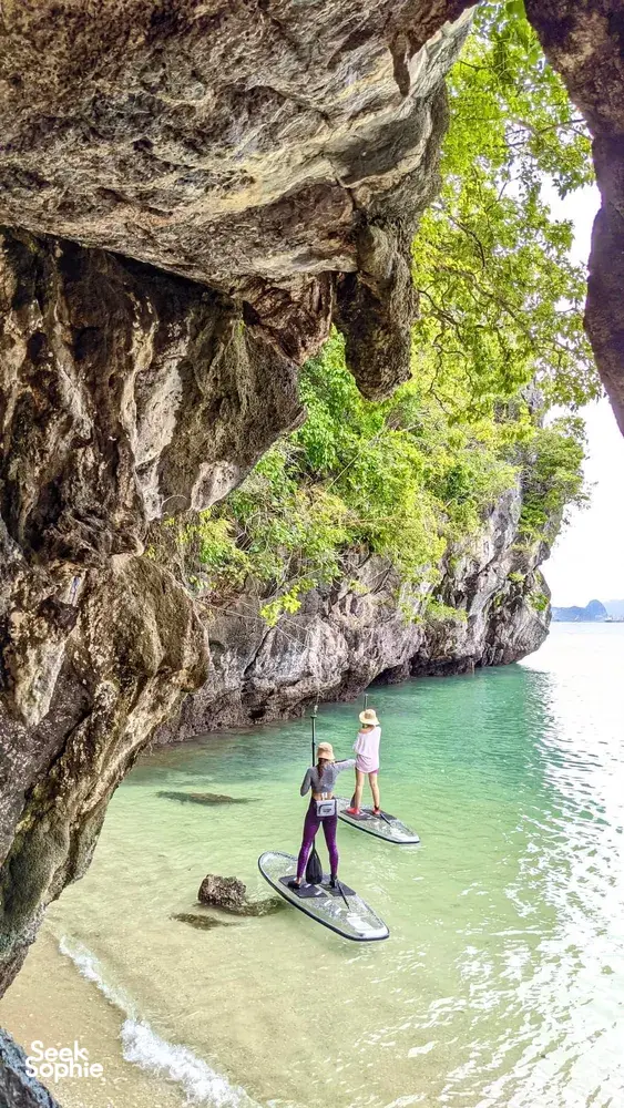 SUP paddlers float beneath a limestone cave wall at Tanjung Rhu, Langkawi, steering through a sheltered turquoise inlet