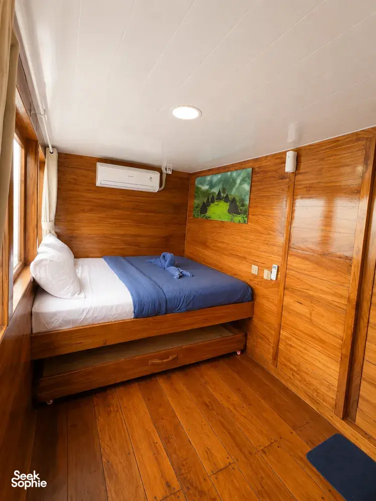 Corner wooden phinisi cabin with queen bed, curtain window and wall artwork on a shared Komodo cruise from Labuan Bajo