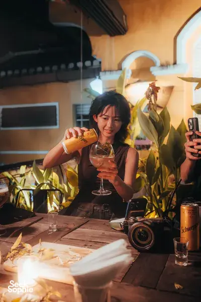 Woman pours a citrus cocktail from a can at a convivial tasting table, with phones up and lantern light in Hoi An