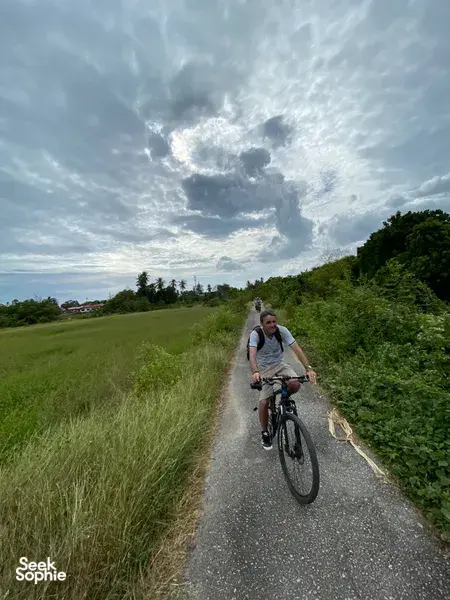 Cyclist pedals along a narrow rural road in Langkawi, past tall grass and hedges under bright, dramatic clouds