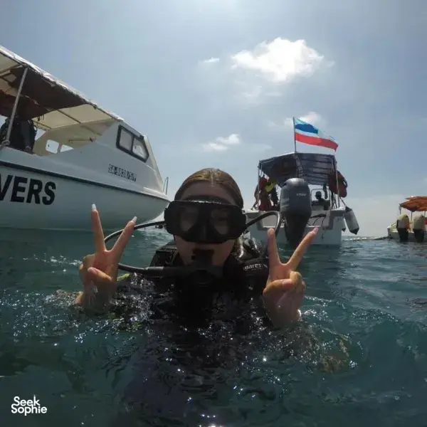 Seorang penyelam di atas bot di perairan Semporna, memakai topeng snorkel dan sut selam, menunjukkan tanda damai sebelum selaman terumbu bersama Bigfin Divers