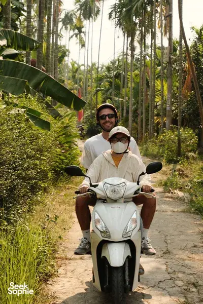 Helmeted motorbike riders smiling on a quiet path through palm trees in Hoi An during the early-morning trades tour