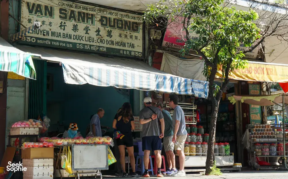 在胡志明市一家越南街头市场摊位前浏览商品的旅行团游客,周围摆满了罐装货品,头顶是遮阳篷