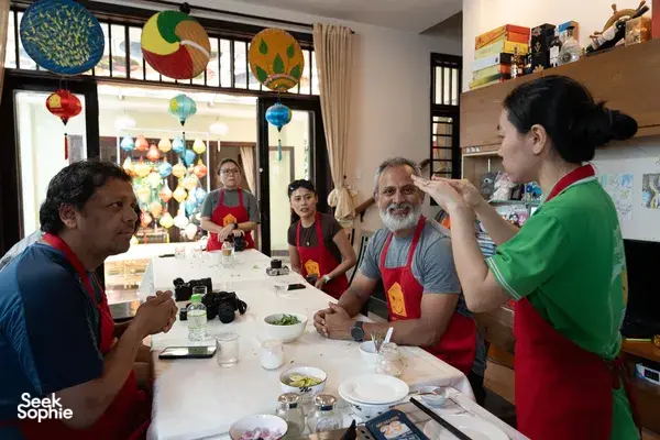 Hoi An cooking class with guests in red aprons preparing ingredients at a table while a host teaches beside colourful lanterns