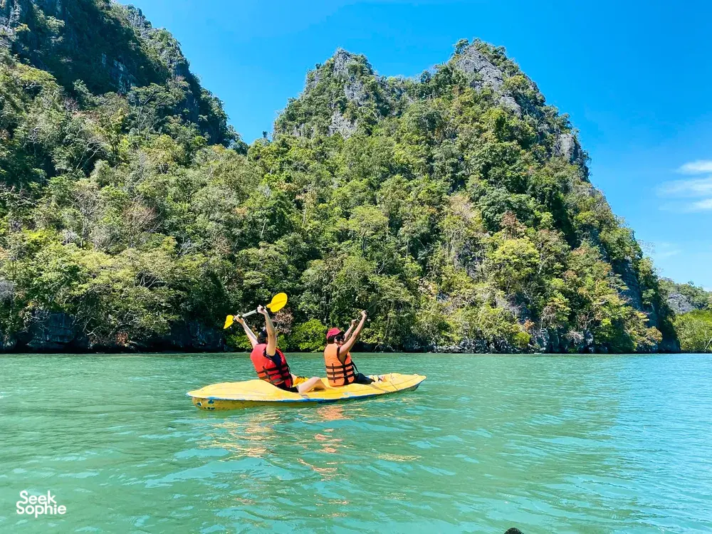 Two people kayaking across clear green water beside forested limestone cliffs in Langkawi during a Kilim Geoforest nature tour