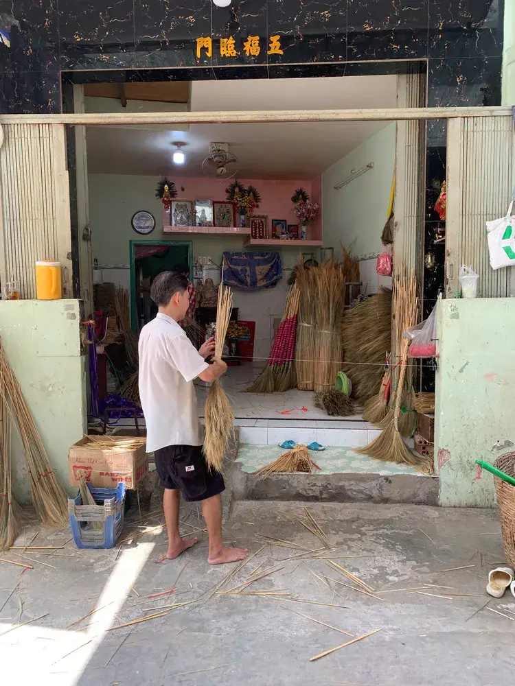 Saigon craft workshop doorway with thatched bundles and floor mats hanging, an artisan holding straw while preparing bundles inside