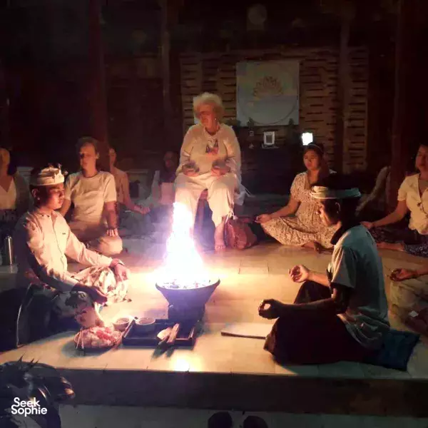 Meditation group seated around a blazing fire during an ADDA Yoga Balinese ritual, with offerings on low floor mats in Canggu