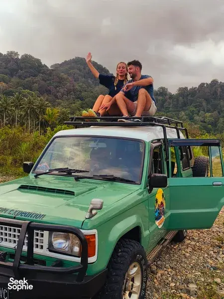 Green 4x4 on a gravel track with two travellers sitting on the bonnet, overlooking lush forested hills in Indonesia