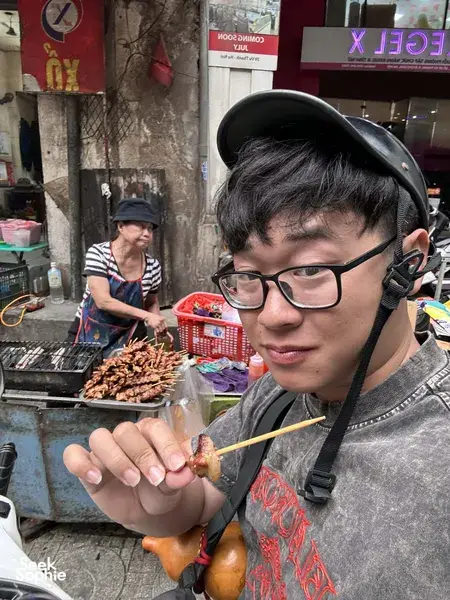 Tour participant wearing a headset eating a grilled skewer at a busy Hanoi street stall, with the grill and ingredients behind