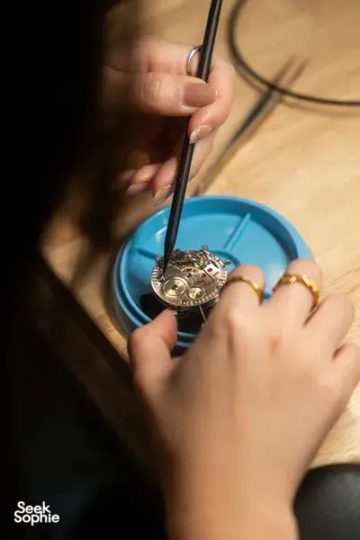 Hands assembling a watch movement with precision tools on a wooden bench, parts in a blue tray — watchmaking workshops in Singapore